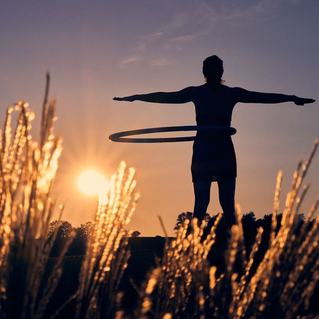 Frau mit Hula Hoop Reifen vor dem Sonnenuntergang, im Vordergund stehen Gräser.